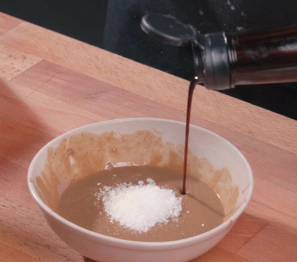 Ein Strahl dunkler Sojasauce wird in eine weiße Keramikschale mit dicker Sesampaste, einem Häufchen weißem Zucker und Essig gegossen.