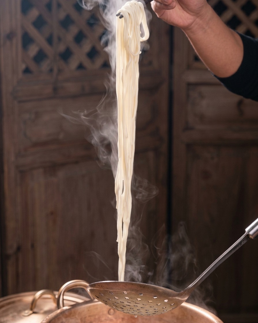 Ein Bündel dampfender weißer Nudeln wird mit dunklen Stäbchen und einem Schaumlöffel aus einem Kupfertopf gehoben.