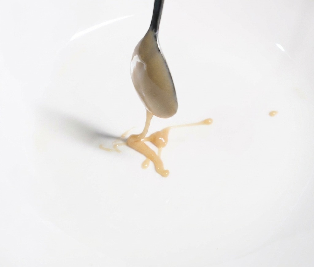 Ein Löffel lässt cremige, hellbraune Sesampaste in eine weiße Schüssel fließen.