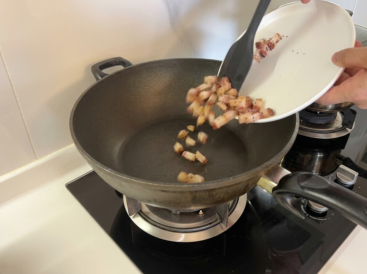 Gewürfelter roher Guanciale wird von einem weißen Teller mit einem Spatel in einen trockenen, unebheizten Wok gegeben.