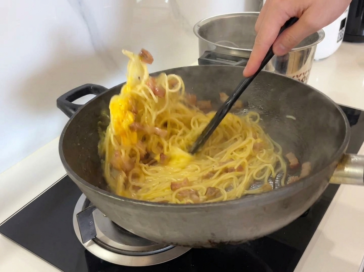 Spaghetti, Guanciale und die gelbe Eiermischung werden mit einem schwarzen Spatel kräftig im Wok gerührt.
