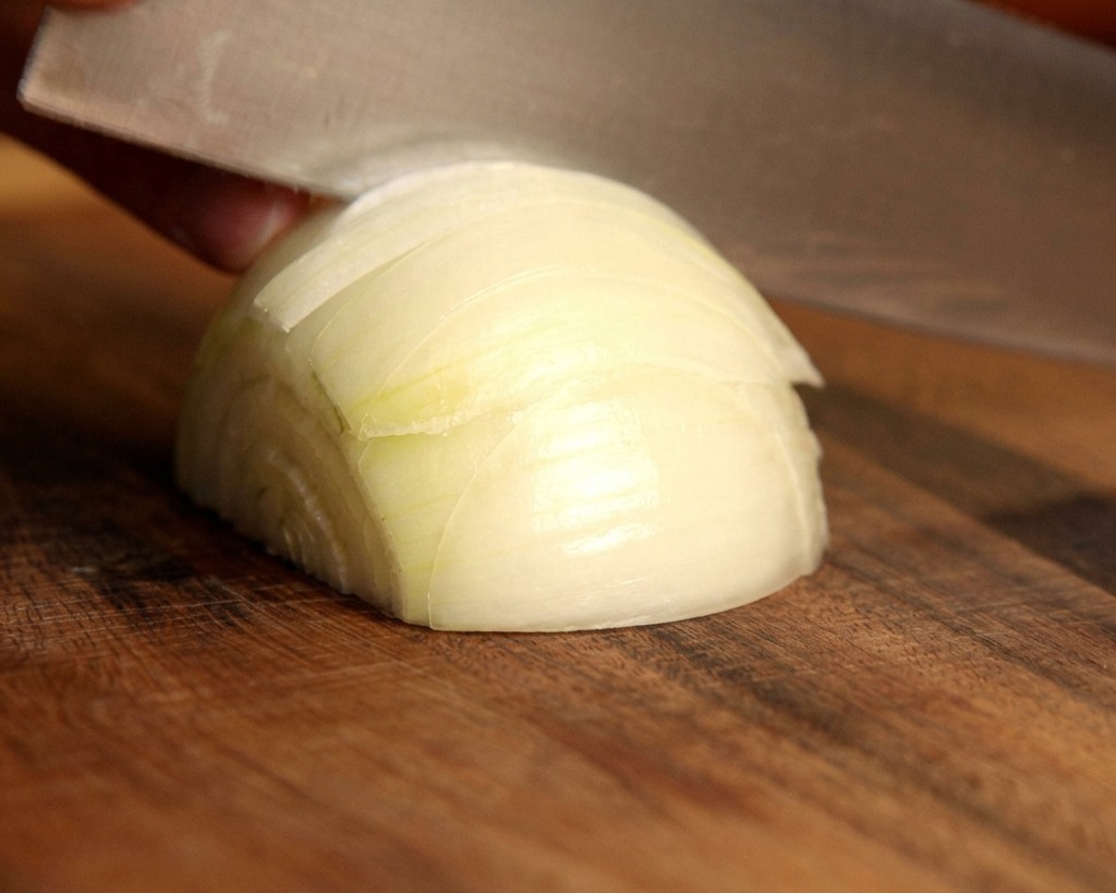 Ein scharfes Messer schneidet eine frische weiße Zwiebel auf einem Holzschneidebrett in dünne Spalten.