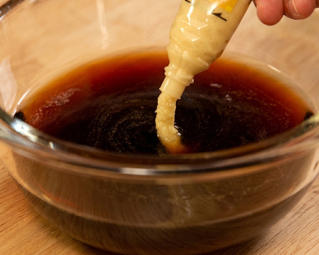 Frisch geriebene Ingwerpaste wird aus einer Tube in eine dunkle Sauce auf Sojabasis in einer Glasschüssel gedrückt.