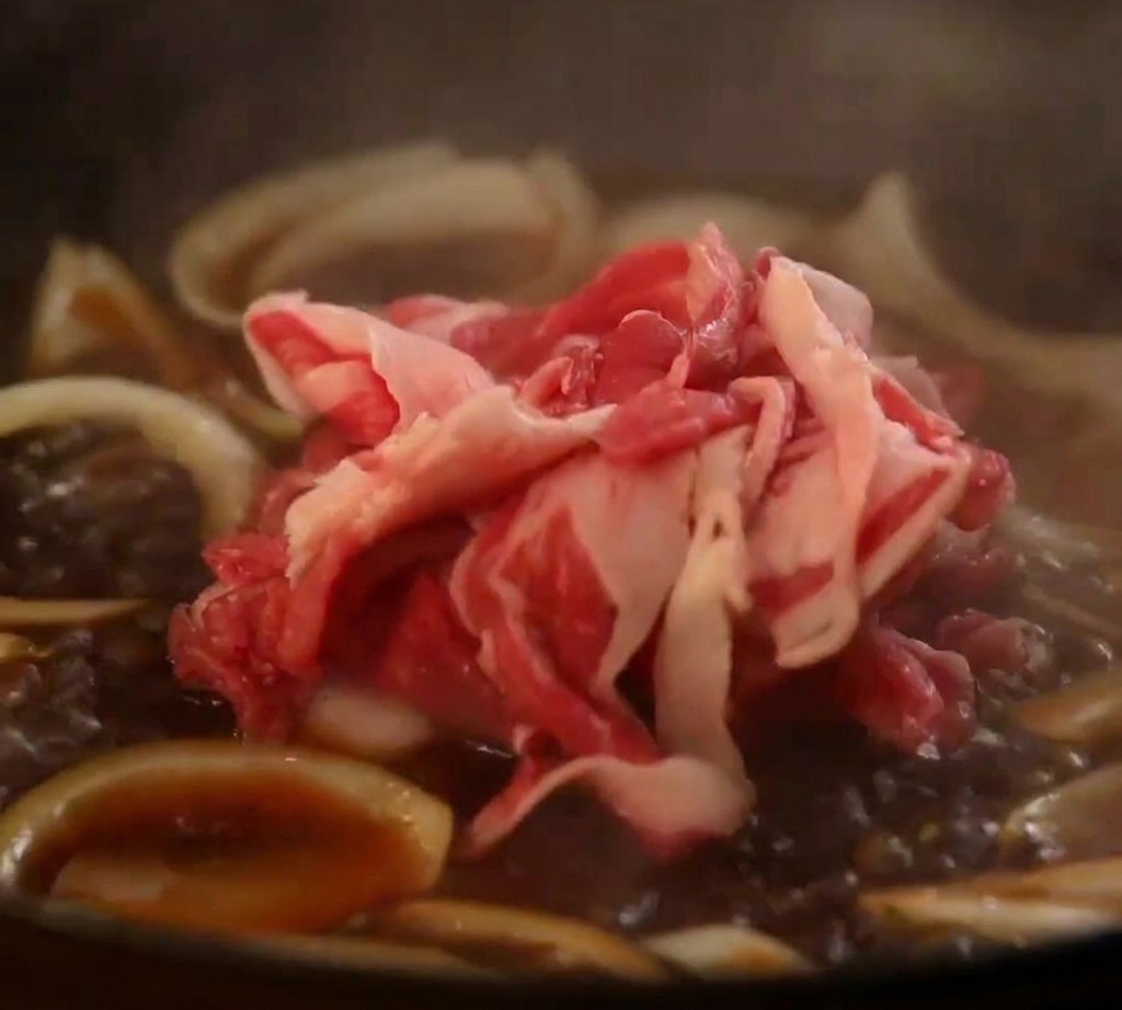 Rohe Rinderbauchscheiben werden in eine köchelnde Pfanne mit Zwiebeln und japanischer herzhafter Sauce gegeben.