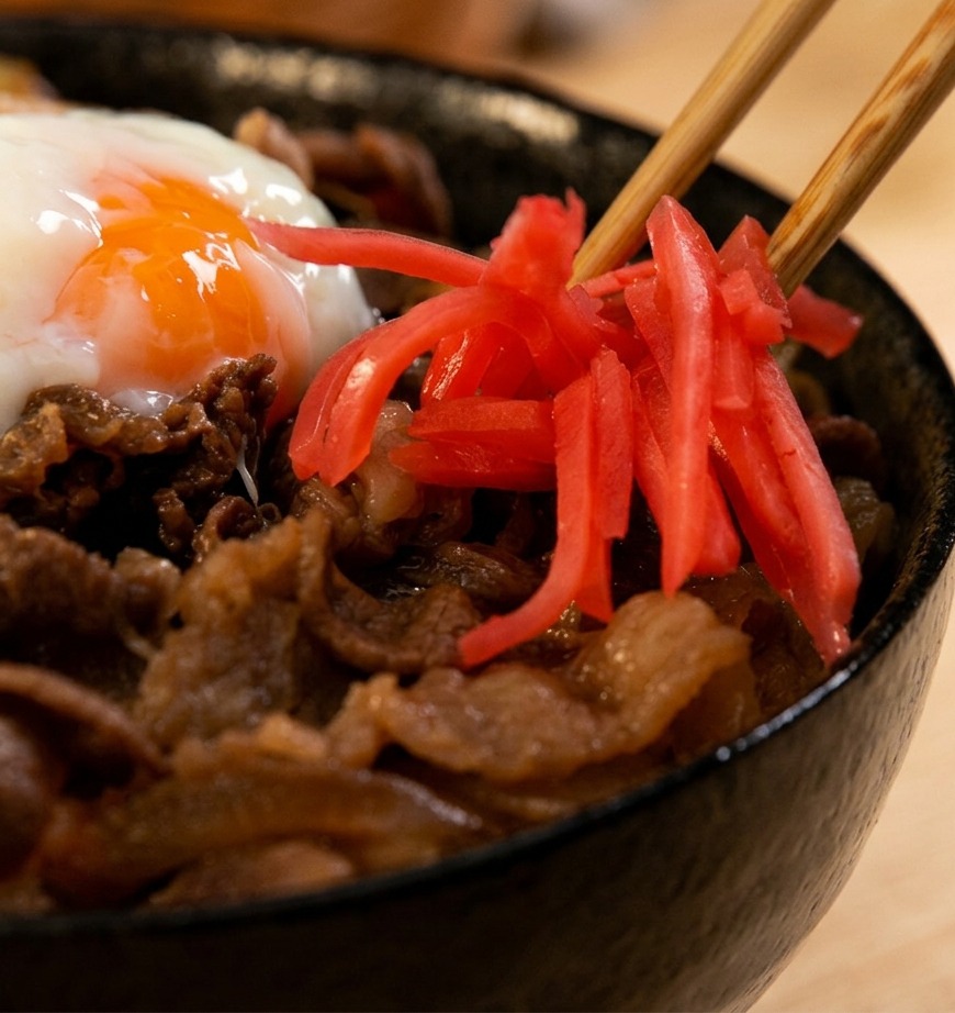 Eine Nahaufnahme von Holzessstäbchen, die leuchtend rote Streifen von eingelegtem Ingwer auf eine dampfende Beef Bowl mit einem weichgekochten Ei legen.
