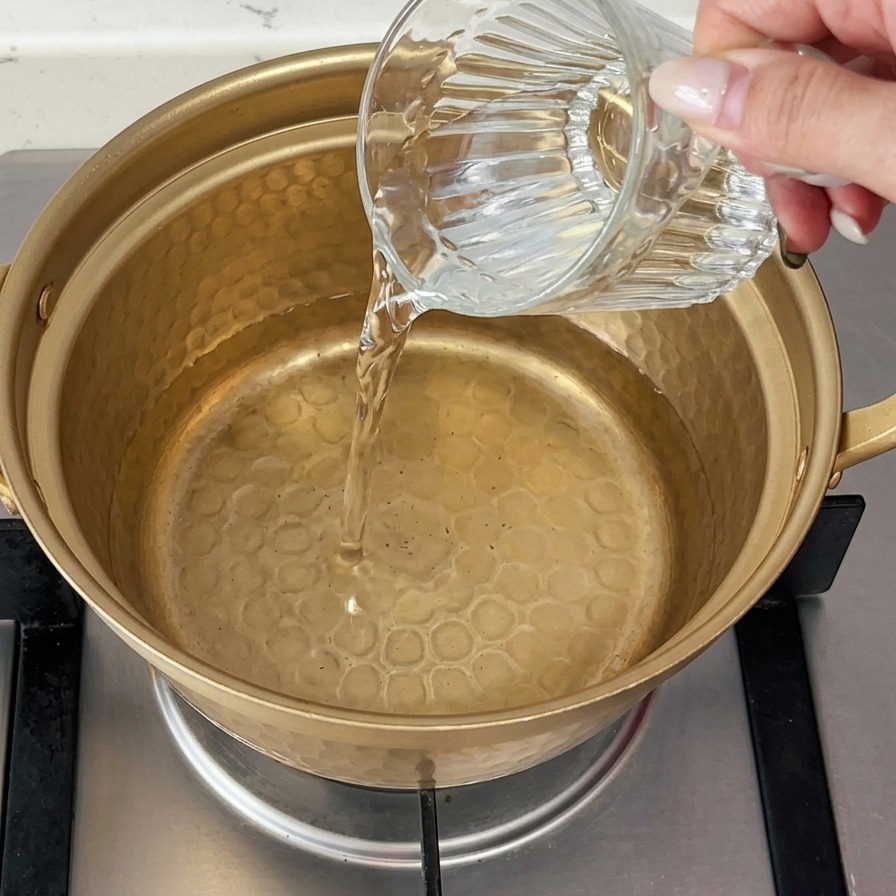 Kaltes Wasser wird aus einem Glas in einen kleinen, goldfarbenen Metalltopf auf einem Gasherd gegossen.