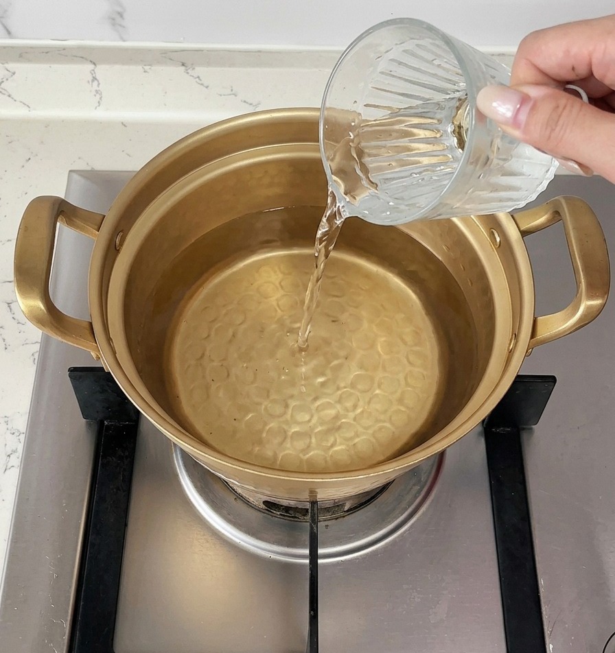 Eine hand gießt eine Tasse kaltes Wasser in einen goldfarbenen Topf mit heißem Wasser auf dem Herd.