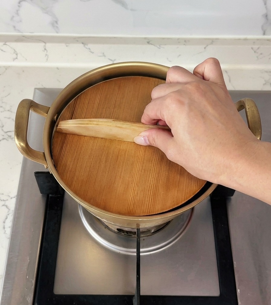 Eine Hand legt einen flachen Holzdeckel auf einen kleinen, goldfarbenen Kochtopf.