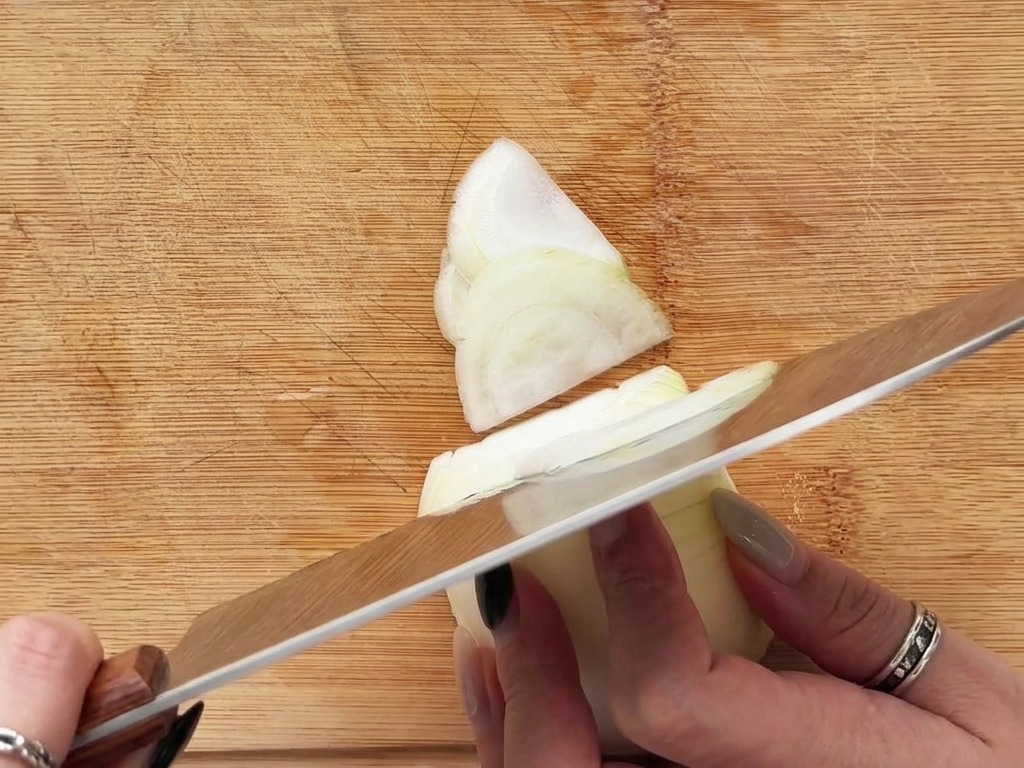 Hände schneiden eine weiße Zwiebel in feine Halbmonde auf einem Holzschneidebrett.