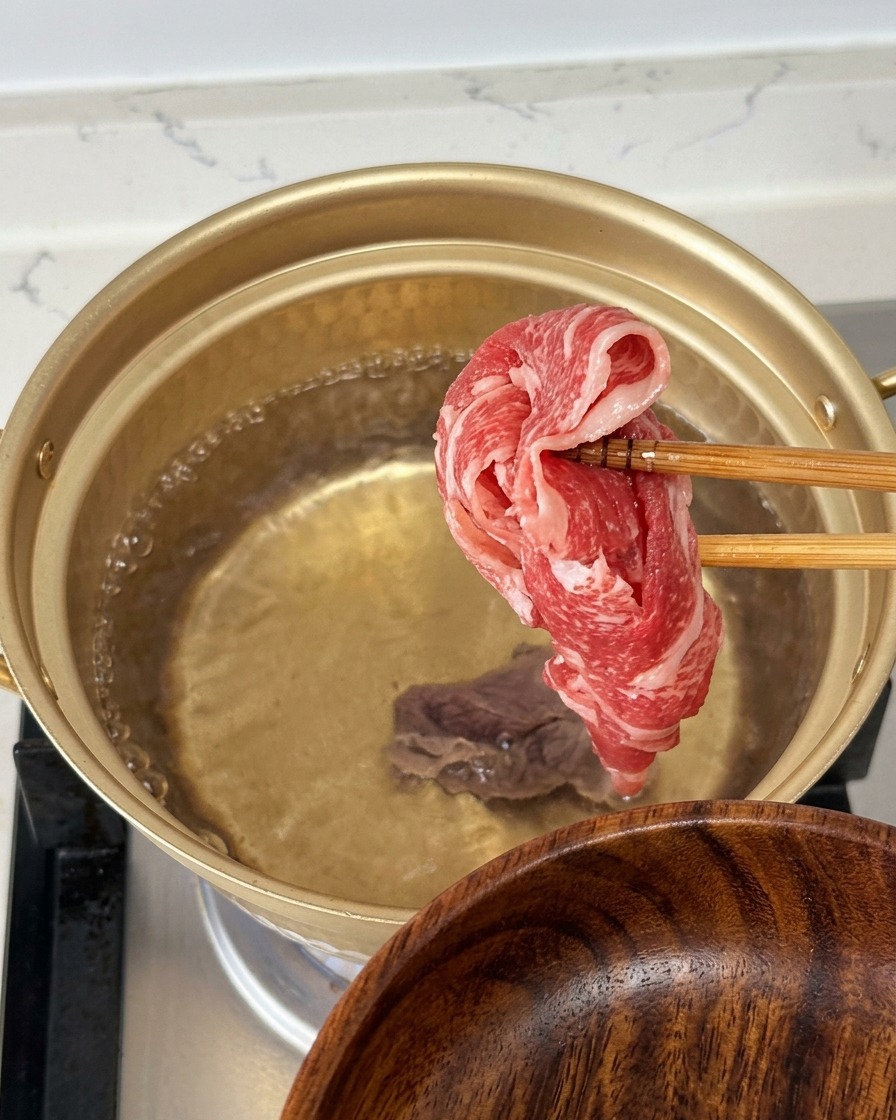 Mit Stäbchen wird eine dünne Scheibe rohes, marmoriertes Rindfleisch über einem kochenden Wassertopf gehalten.