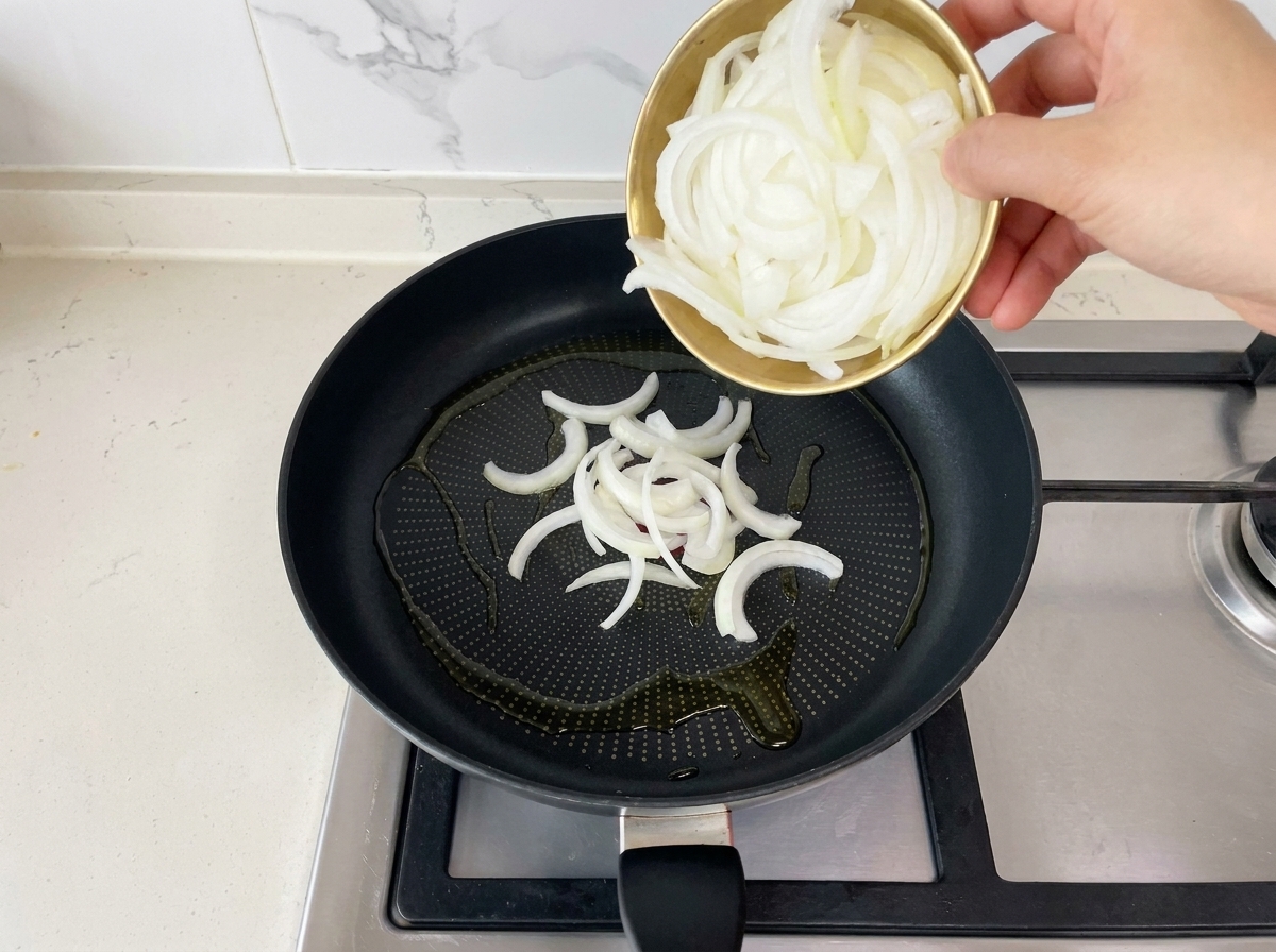 Frisch geschnittene weiße Zwiebeln werden aus einer kleinen Goldschale in eine schwarze Bratpfanne mit heißem Öl gegeben.