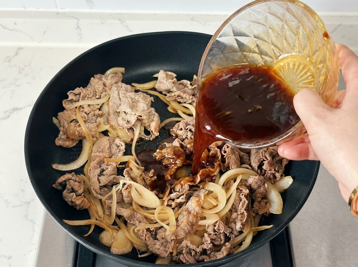 Eine Hand gießt eine dunkle Würzsauce aus einer glasschale über Rindfleisch und Zwiebeln in einer schwarzen Pfanne.