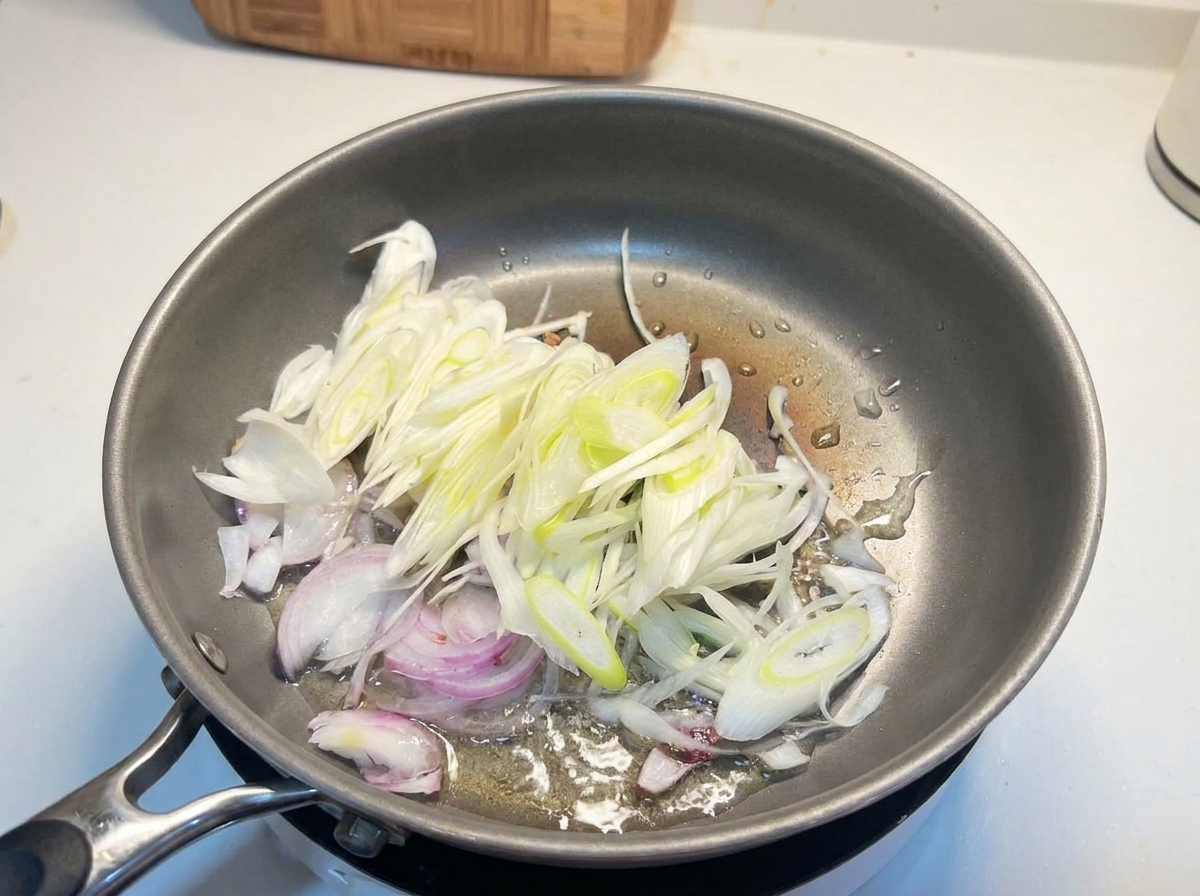 In Streifen geschnittene weiße Zwiebeln und Frühlingszwiebeln braten in heißem Öl in einer Metallpfanne.