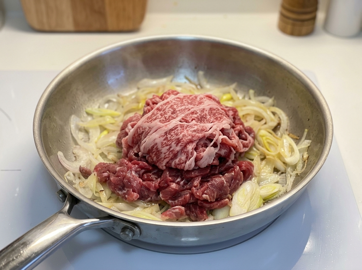 Ein Haufen rohes, dünn geschnittenes Rindfleisch liegt auf angebratenen Zwiebeln in einer Metallpfanne.