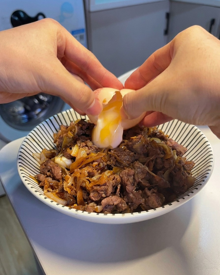 Zwei Hände schlagen ein Onsen-Ei auf, sodass ein weiches, flüssiges Eigelb und eiweiß auf eine dampfende Rindfleisch-Reis-Bowl gleiten.