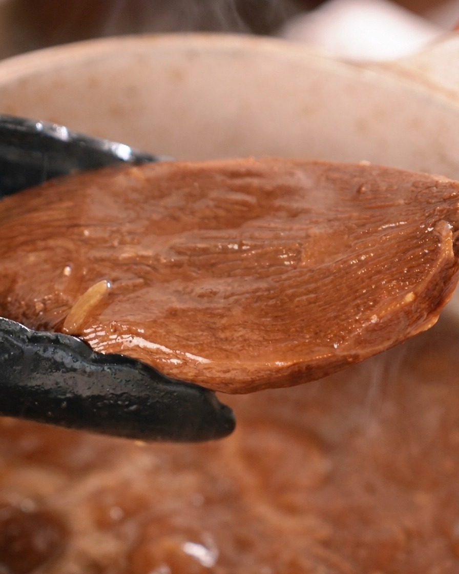Ein dickes Stück geschmorter Rinderzunge wird mit einer schwarzen Küchenzange aus einem Topf mit dunkler Sauce gehoben.