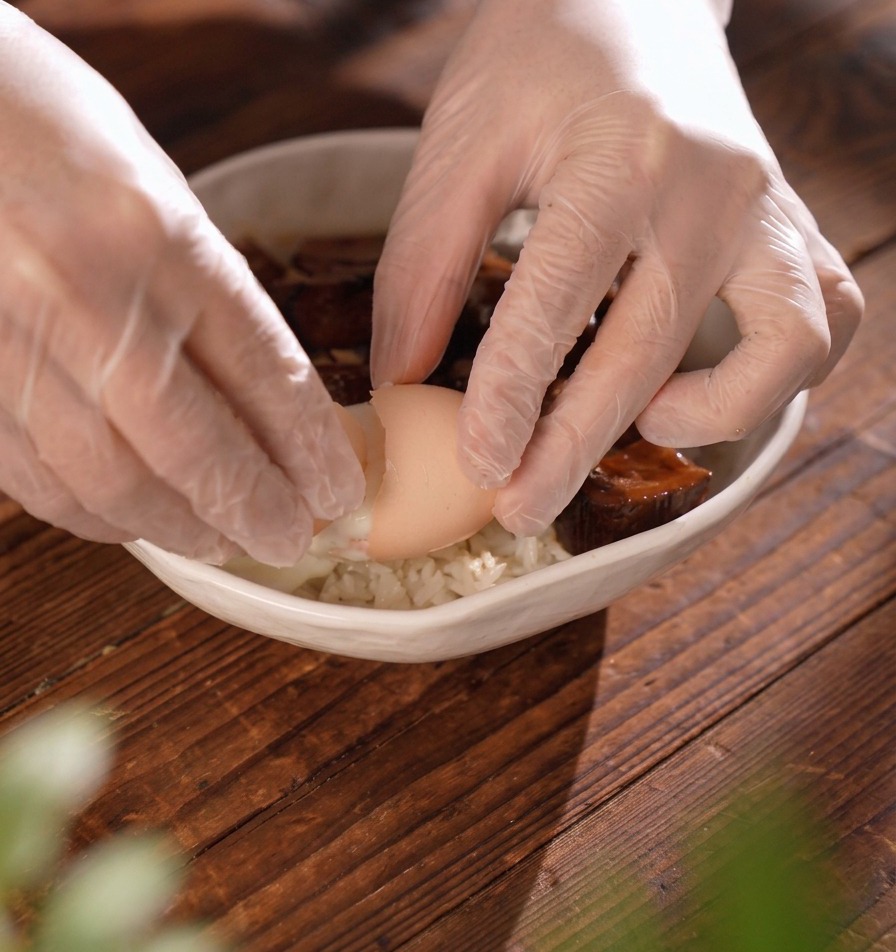 Behandschuhte Hände brechen behutsam eine weiche Eierschale auf, um ein Onsen-Ei zu dem Fleisch in der Reisschale gleiten zu lassen.