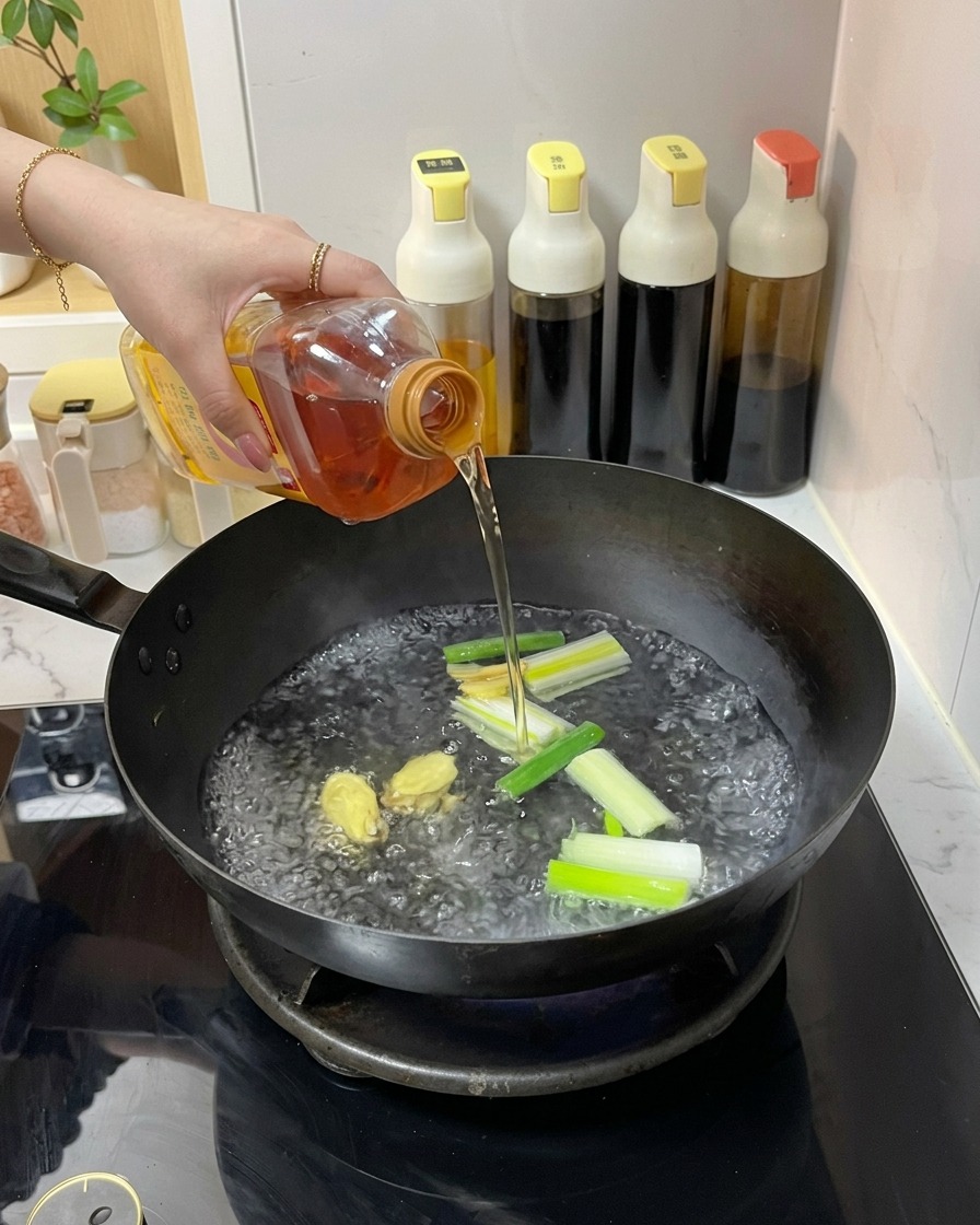Eine Person gießt eine klare bernsteinfarbene Flüssigkeit aus einer Plastikflasche in einen Wok mit kochendem Wasser, Ingwer und Frühlingszwiebeln.