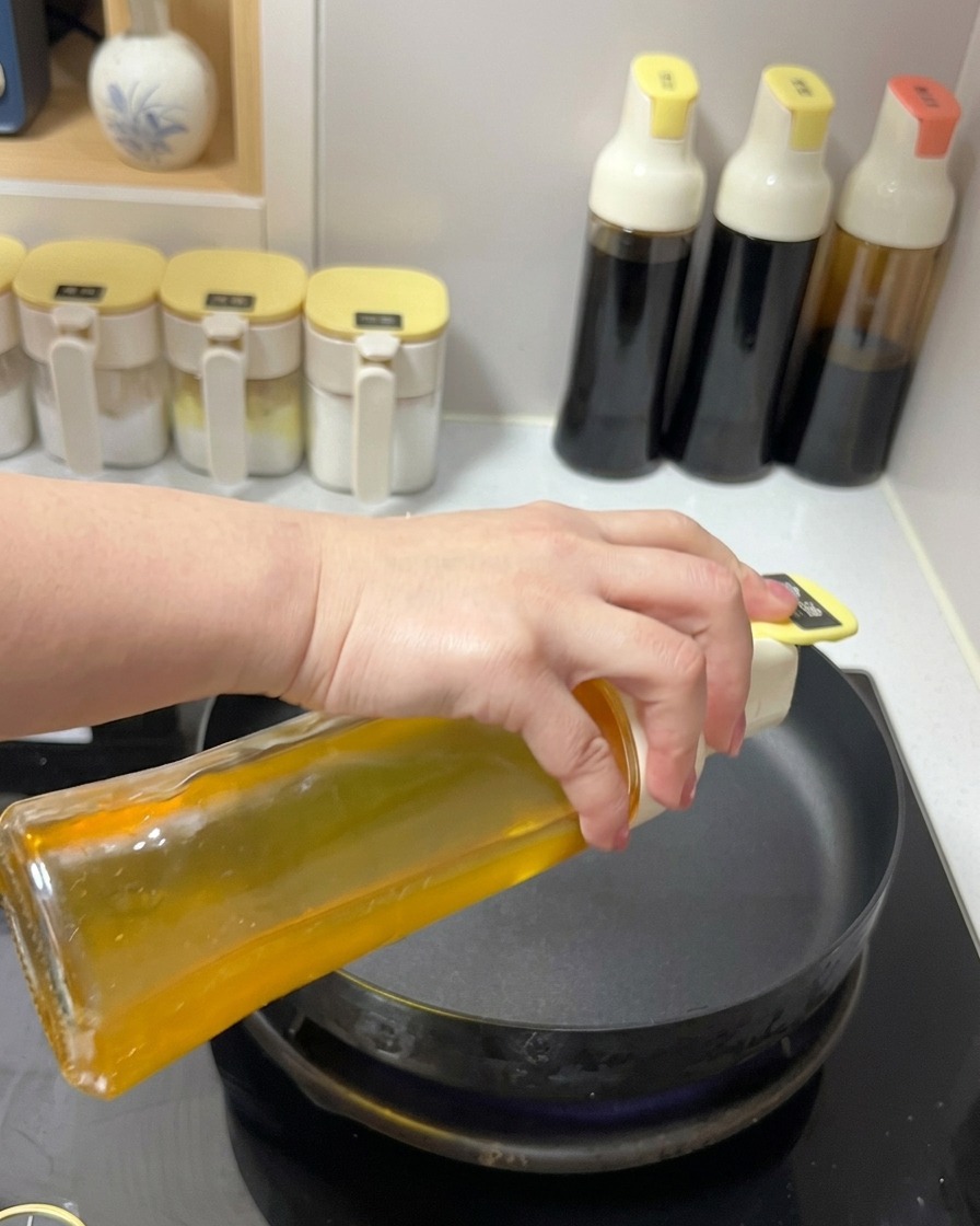 Klares Pflanzenöl wird aus einem Glasspender in eine schwarze Bratpfanne auf einem Herd gegossen.