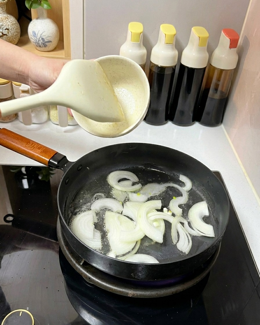Weiße Zwiebelscheiben werden mit einem weißen Pfannenwender in einer Pfanne mit Öl geschwenkt.