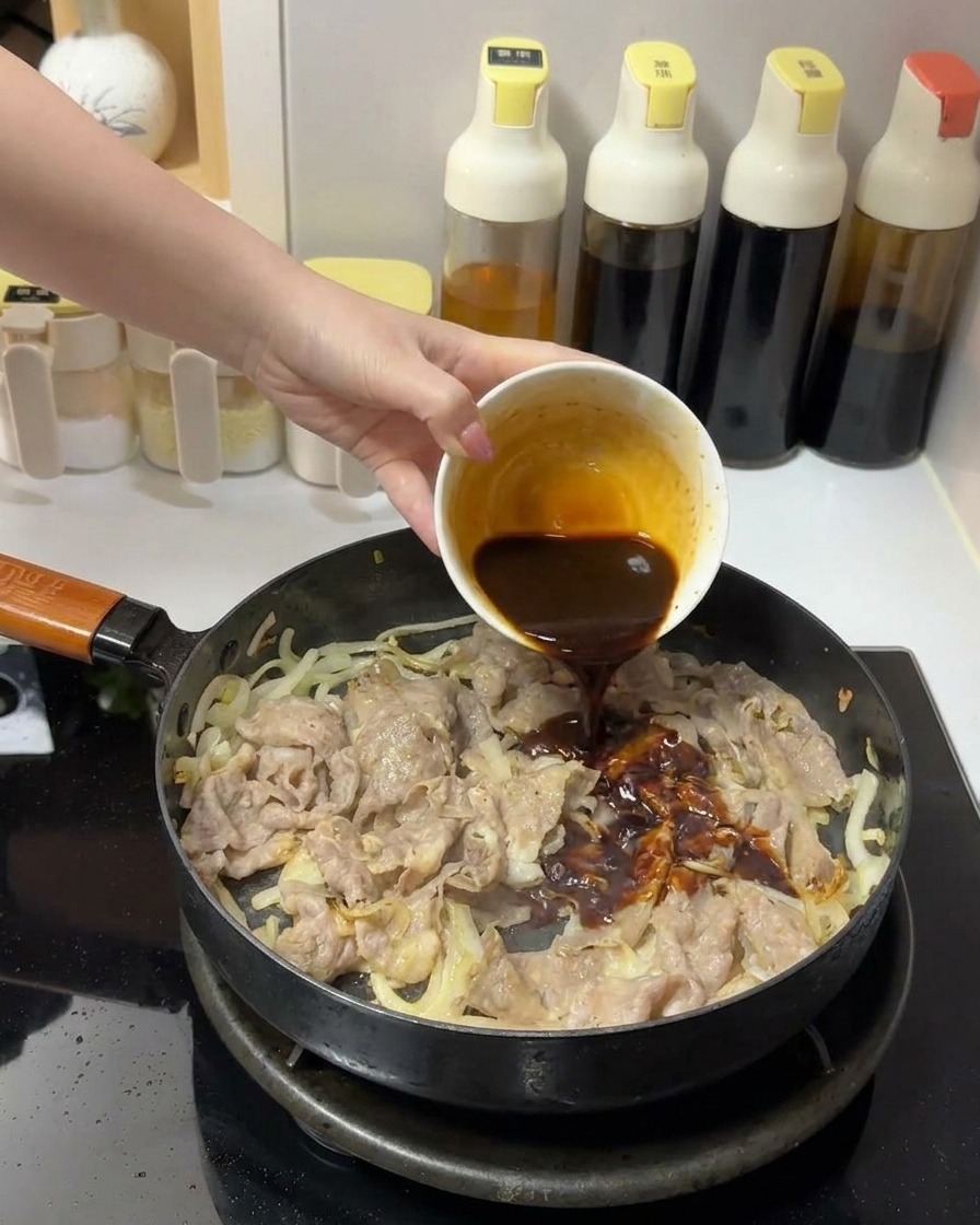 Eine dicke, dunkelbraune Soße wird aus einer kleinen weißen Schale über das Fleisch und die Zwiebeln in einer Bratpfanne gegossen.