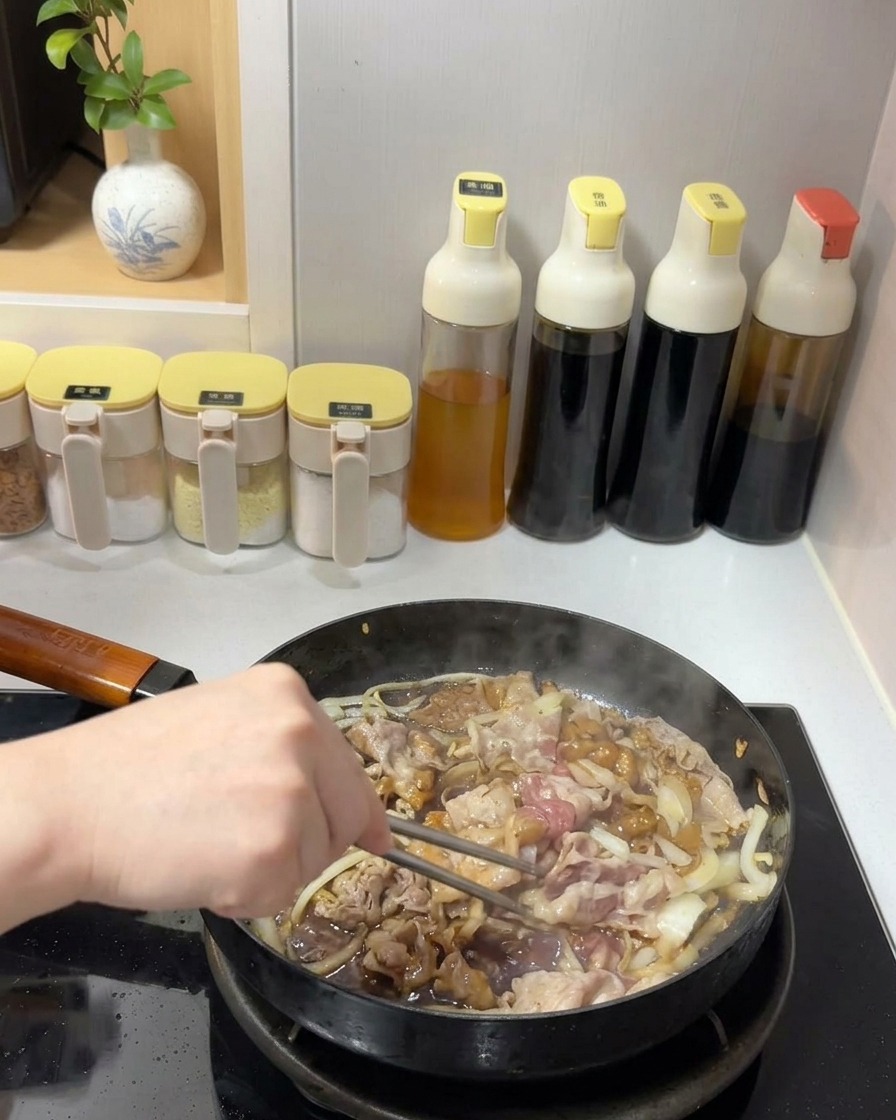 Rindfleisch und Zwiebeln werden in einer dunklen, glänzenden Soße in einer Pfanne mit Metallstäbchen durchgeschwenkt.