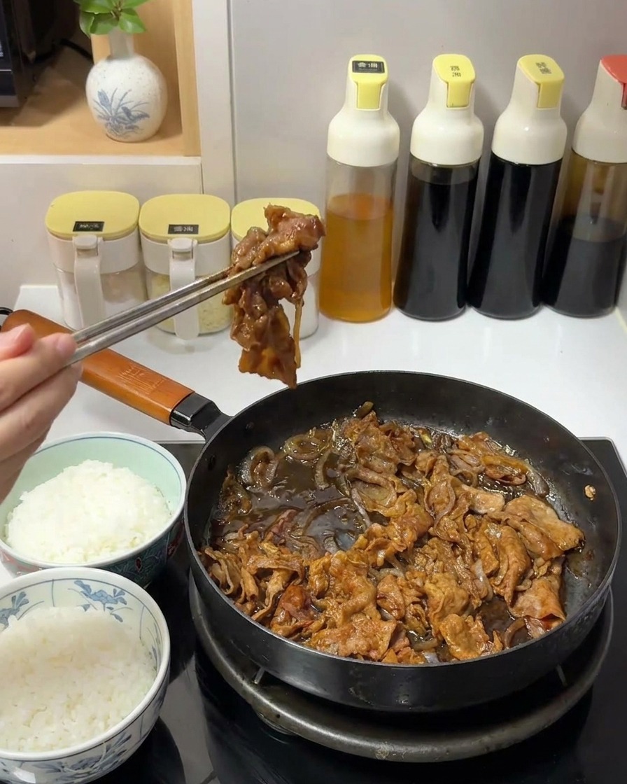 Eine Hand hebt mit Metallstäbchen eine Portion dunkles, saftiges Rindfleisch und Zwiebeln aus einer Pfanne.