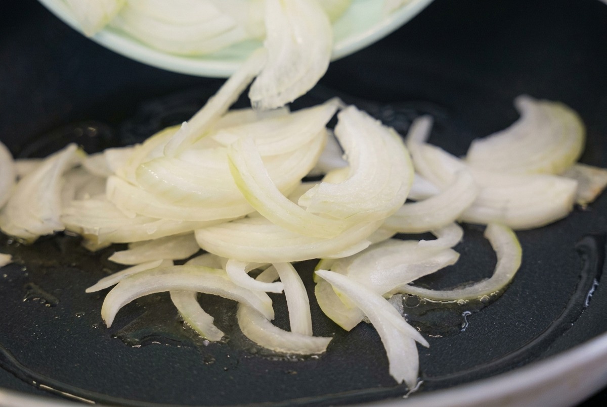Rohe weiße Zwiebelstreifen werden von einem hellgrünen Teller in eine schwarze Bratpfanne mit heißem Öl gegeben.