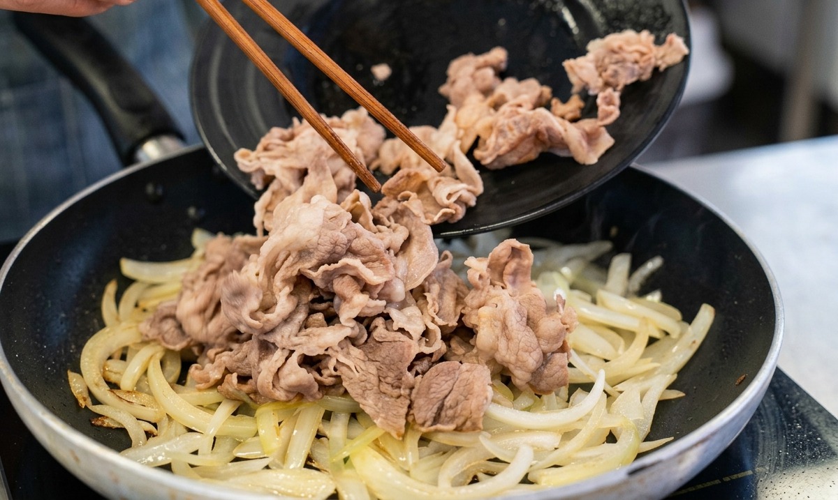 Eine Person lässt blanchierte Rindfleischstreifen mit Holzstäbchen von einem schwarzen Teller in eine Pfanne mit angedünsteten Zwiebeln gleiten.
