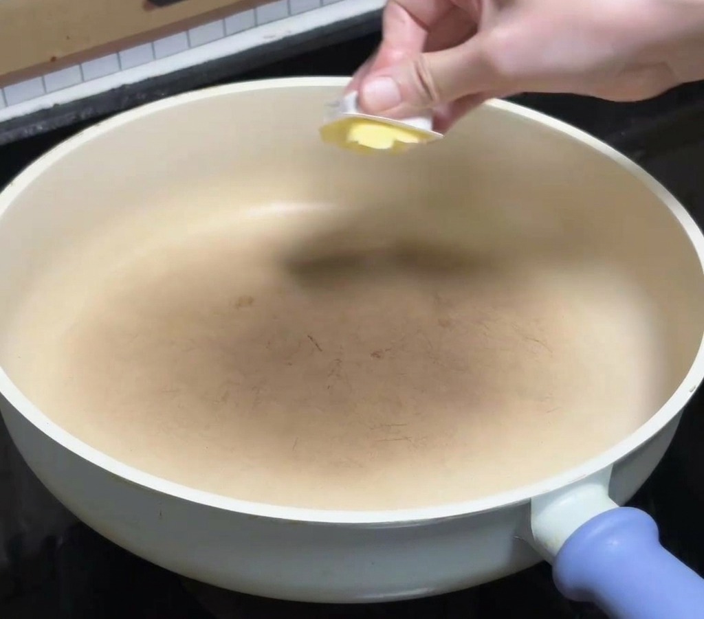 Eine Hand gibt ein Stück gelbe Butter in eine saubere, weiße Keramikpfanne auf dem Herd.