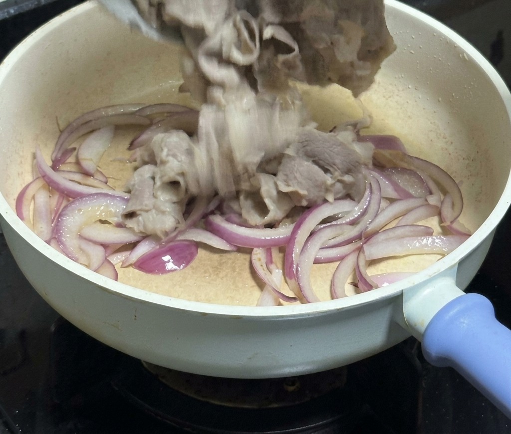Dünn geschnittenes Rindfleisch wird aus einer weißen Schüssel in eine Pfanne mit glasigen roten Zwiebelstreifen gegeben.