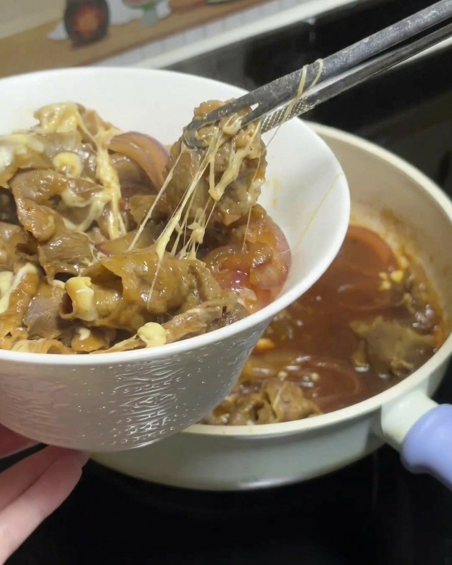 Eine Zange hebt eine Portion Teriyaki-Rindfleisch mit geschmolzenem Käse aus der Pfanne, wobei lange Käsefäden zu sehen sind, und platziert sie auf einer Schüssel mit weißem Reis.