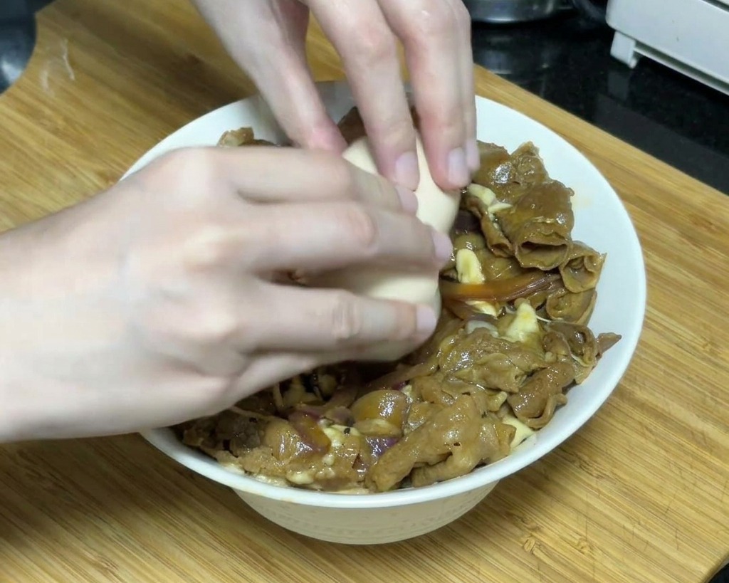 Zwei Hände schlagen vorsichtig ein weich gegartes Onsen-Ei über einer Schüssel mit Teriyaki-Rindfleisch und geschmolzenem Käse auf.