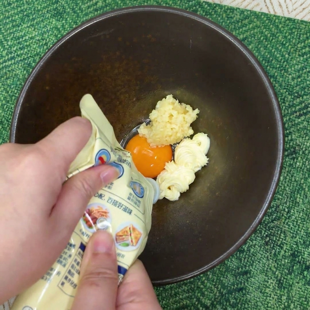 Hände drücken Mayonnaise aus einer Tube auf ein Eigelb und Knoblauch in einer dunklen Keramikschüssel.