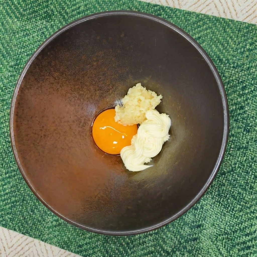 Dunkle Schüssel mit Eigelb, Mayonnaise und Knoblauch auf einem grünen Platzset.
