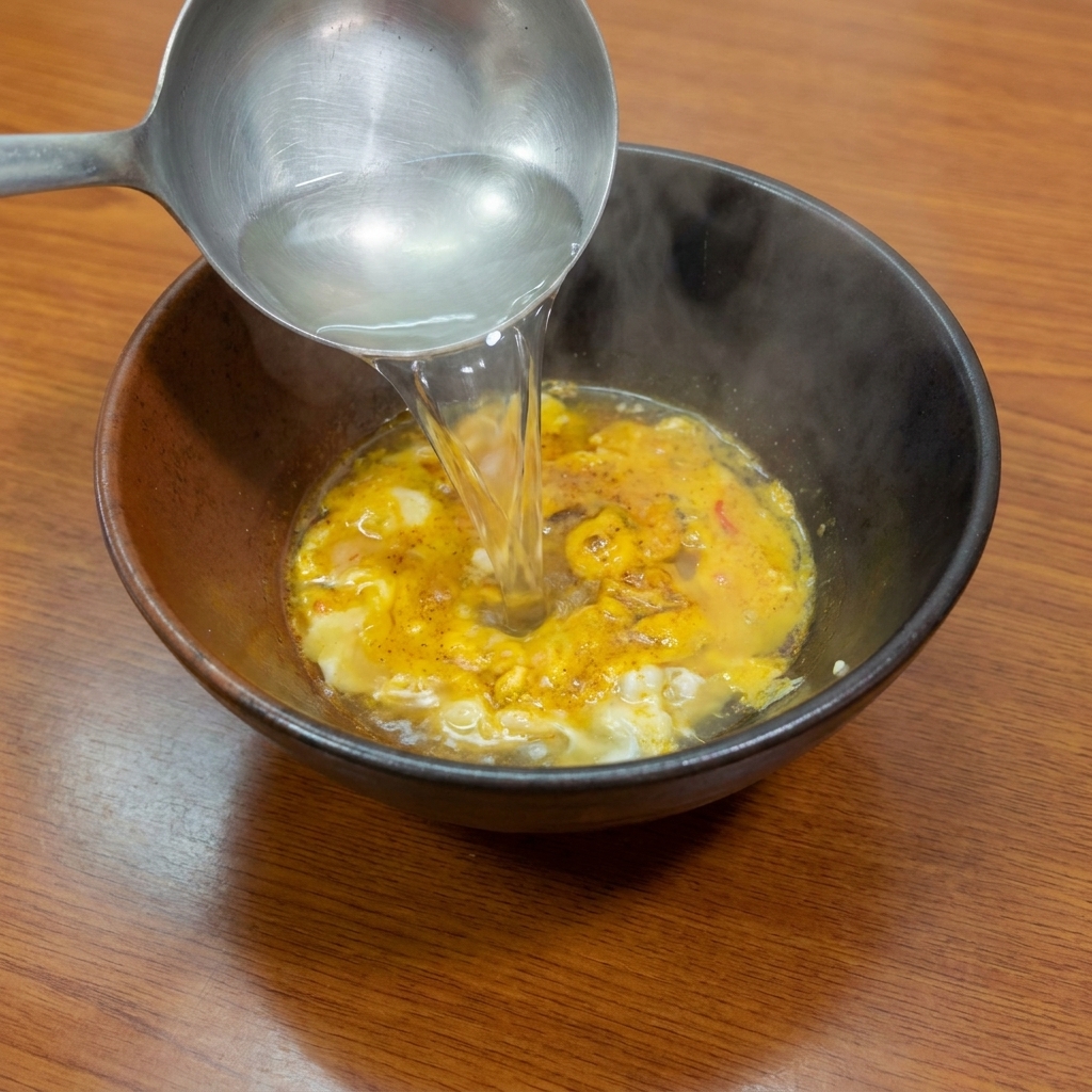Eine Schöpfkelle gießt kochendes Wasser über die gelbe Gewürzpaste in einer dunklen Schüssel.