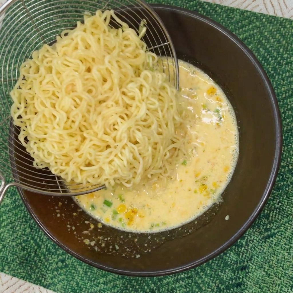 Ein Metallsieb gibt gekochte Instant-Nudeln in eine Schüssel mit cremiger gelber Brühe.