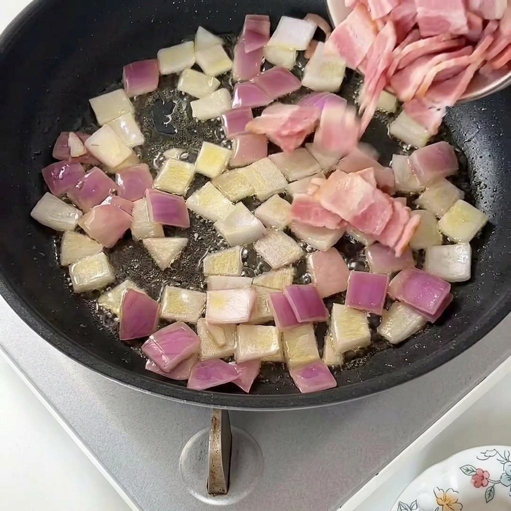 Rohe Speckstücke werden aus einer kleinen Schüssel in die Pfanne zu den Zwiebeln gegeben.