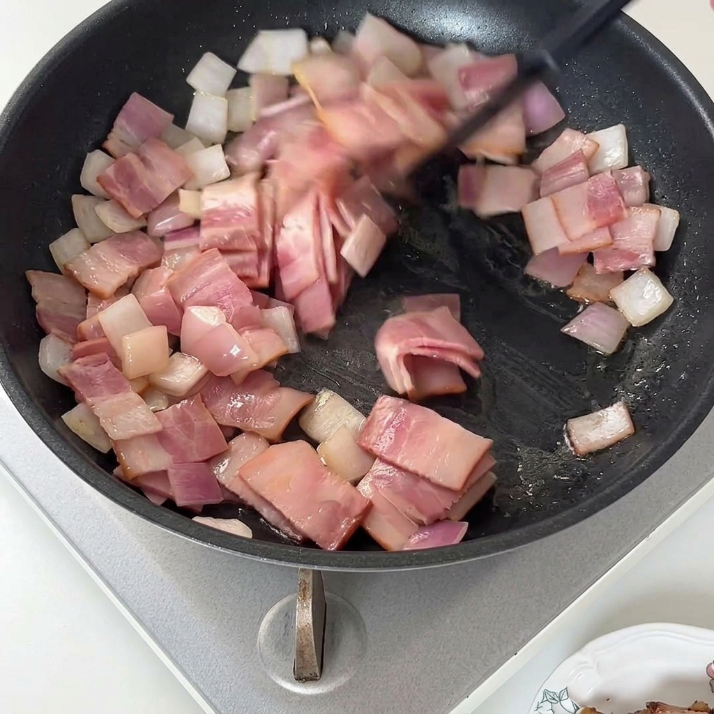 Ein Pfannenwender rührt gebratene Speckstücke und Zwiebeln in einer Pfanne um.