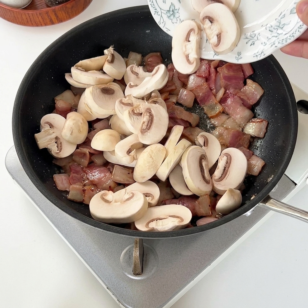 Geschnittene Champignons werden zu dem gebratenen Speck und den Zwiebeln in die Pfanne gegeben.