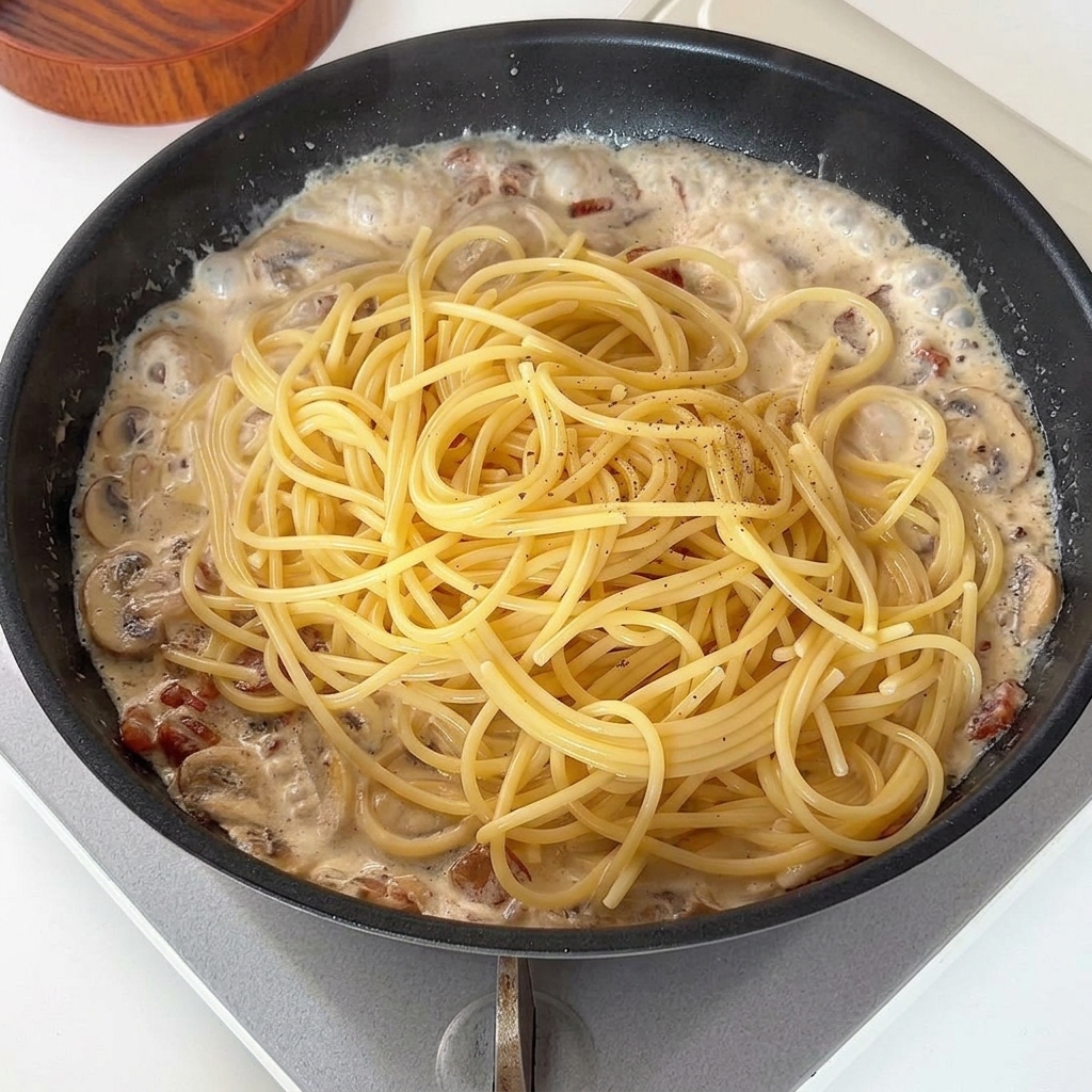Gekochte Spaghetti werden auf die cremige Sauce in der Pfanne gegeben und mit Pfeffer bestreut.
