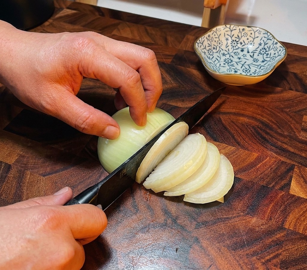 Ein Kochmesser schneidet eine weiße Zwiebel auf einem dunklen Holzschneidebrett in feine Streifen.