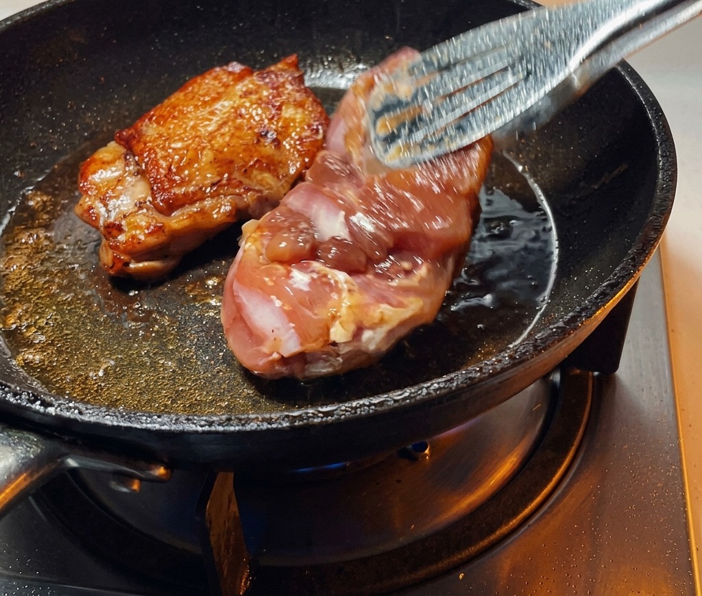 Eine Küchenzange wendet ein angebratenes Stück Hähnchenfleisch in der Pfanne und legt die perfekt gebräunte, knusprige Haut frei.