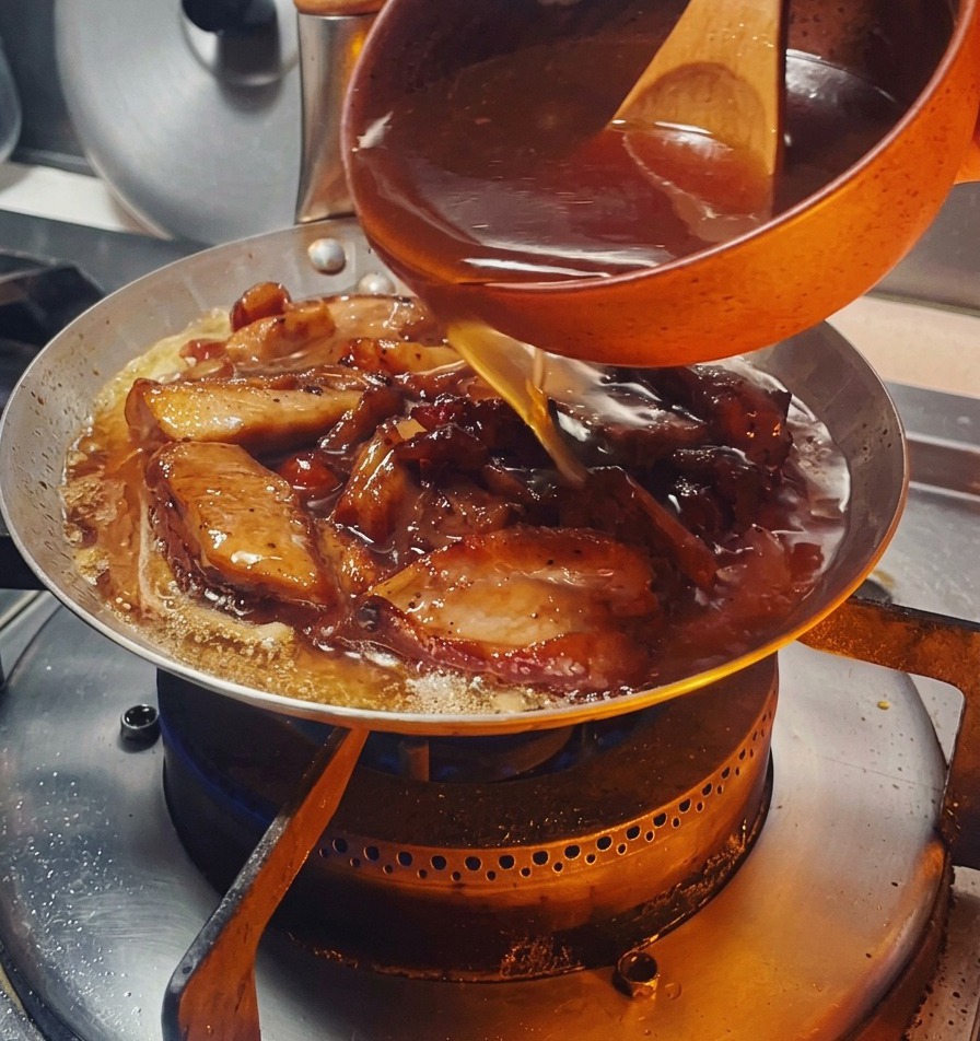 Eine dunkle, herzhafte Sauce wird aus einer kleinen orangefarbenen Schale über das gebräunte Hähnchen und die Zwiebeln in der Pfanne gegossen.