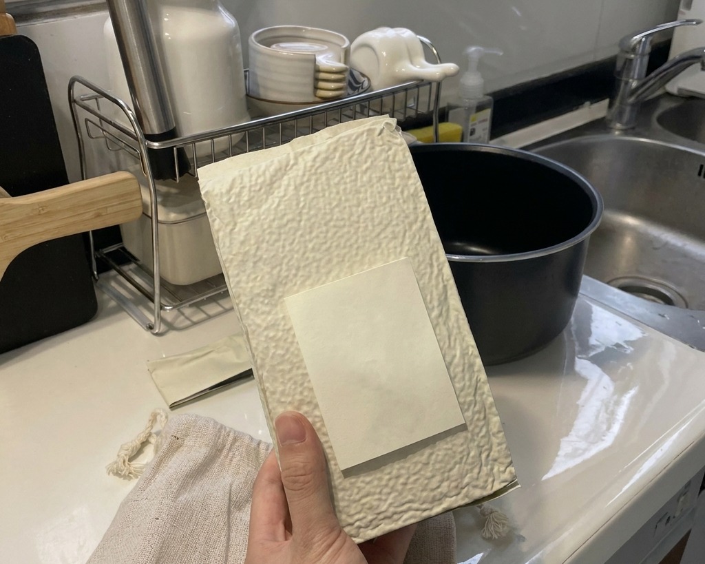 A hand holding a white textured bag, pouring dry white short-grain rice into the black inner pot of a rice cooker on a clean kitchen counter.