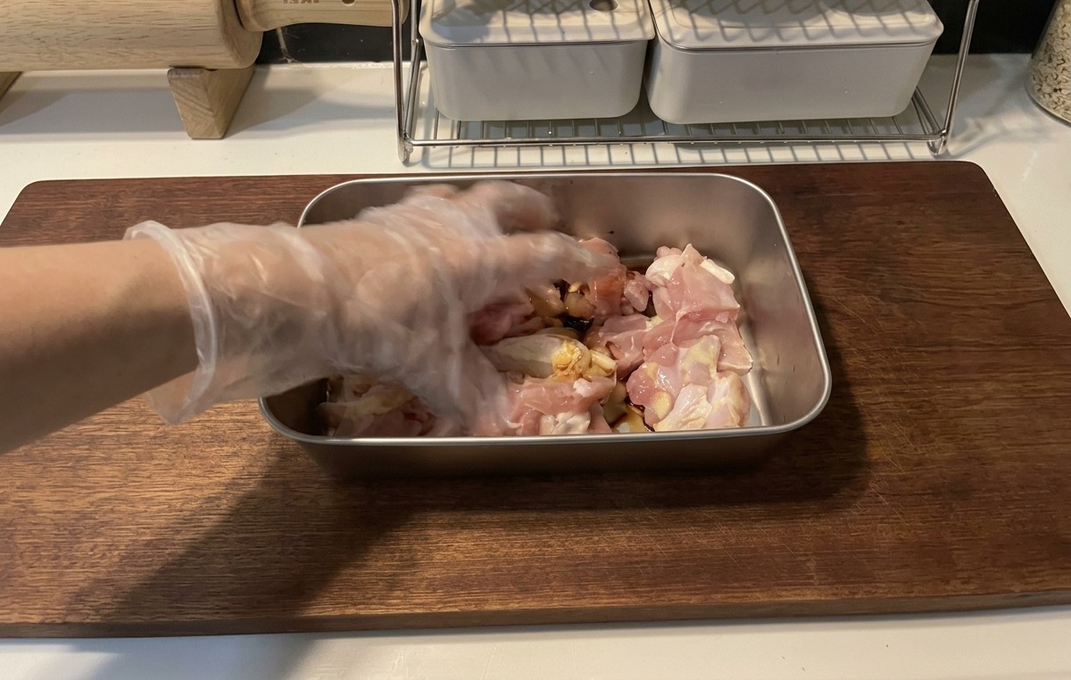 A hand wearing a clear plastic glove mixing raw chicken pieces with a dark soy sauce marinade in a shallow metal tray on a wooden cutting board.
