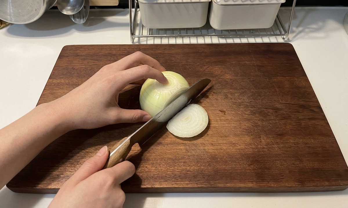 Hands using a wooden-handled knife to slice a white onion into thin crescent strips on a dark wooden cutting board.