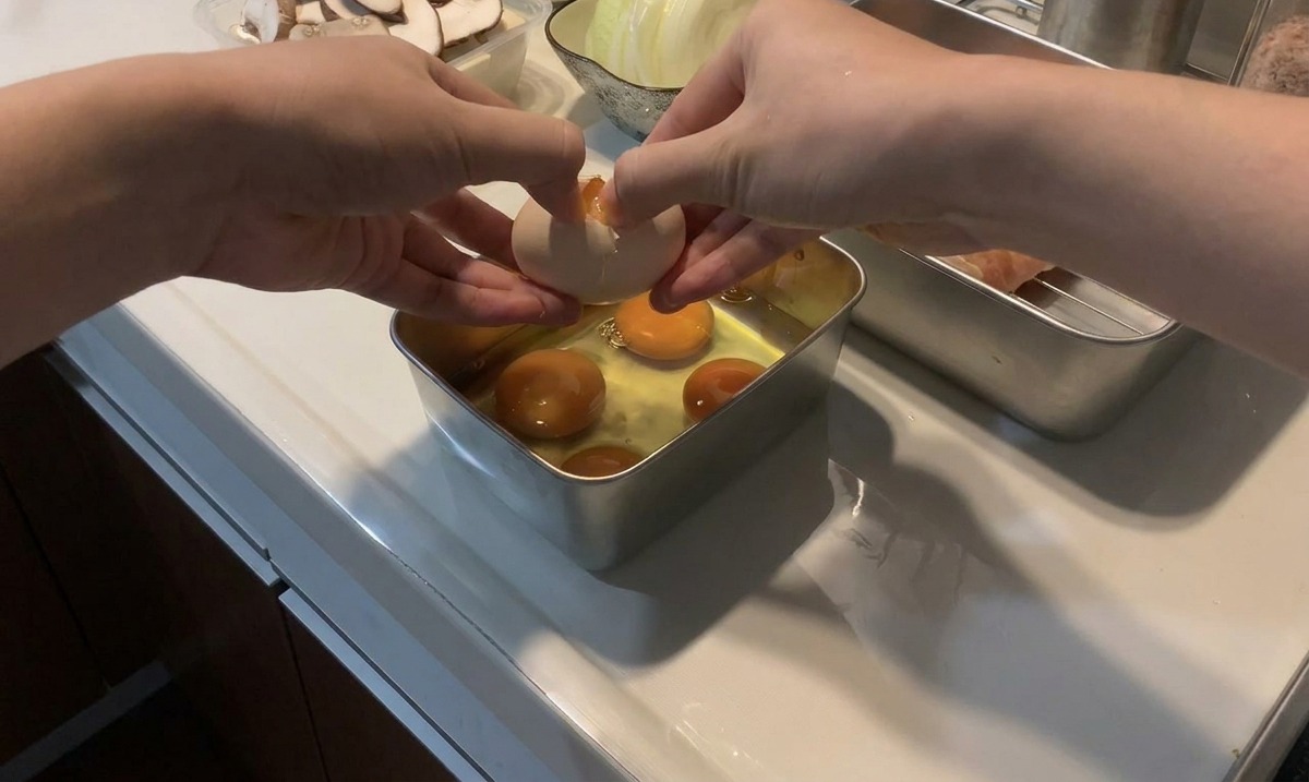 Hands cracking a fresh egg over a metal tray that already contains several uncooked egg yolks and whites.