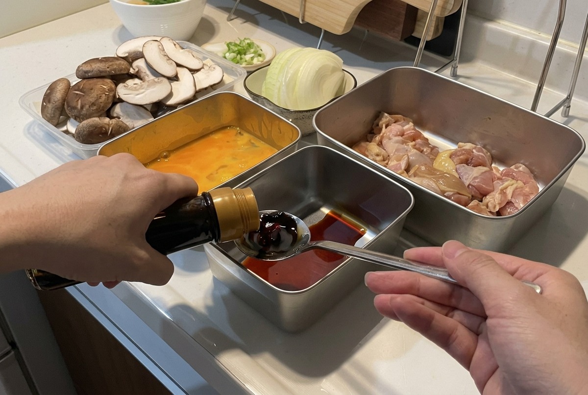 A person pouring dark soy sauce from a bottle into a spoon over a metal container filled with liquid, surrounded by prepped ingredients like mushrooms and sliced onions.