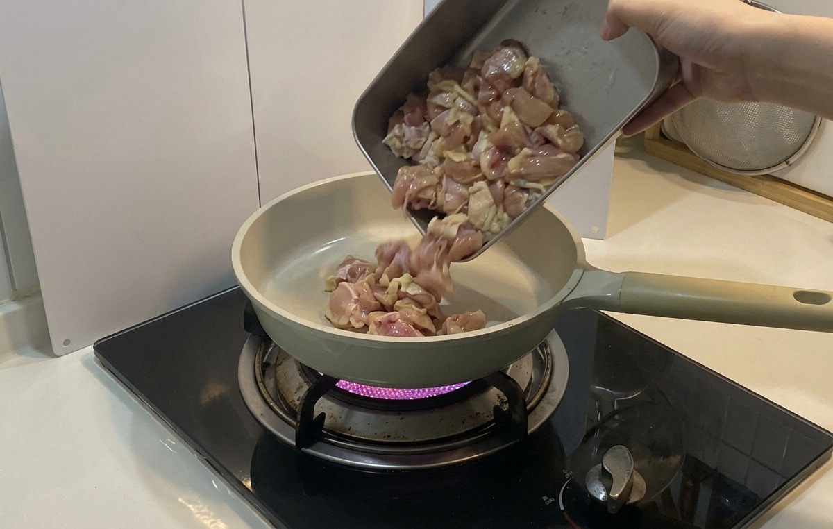 Marinated chicken pieces being poured from a metal tray into a light-colored ceramic frying pan over a purple gas flame.